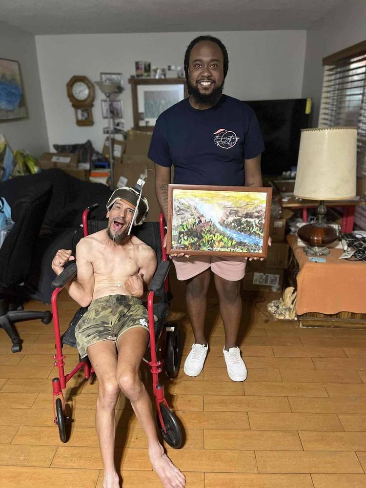 A man in a red wheelchair smiles at home while a caregiver stands beside him holding a framed picture
